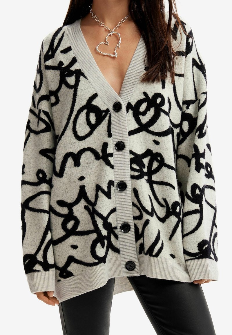 Femme portant un cardigan gris avec des motifs abstraits noirs en forme de script, des boutons noirs sur le devant, associée à un pantalon noir et un collier épais en forme de cœur.