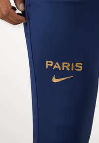 Tamsiai mėlynos sportinės kelnės su "PARIS" ir Nike logotipu auksinės spalvos ant šlaunies. Lygus paviršius ir prigludęs dizainas.