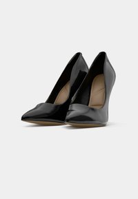 Des chaussures à talons hauts en cuir verni noir avec des pointes effilées et un design élégant, présentant une texture lisse et un intérieur beige subtil.