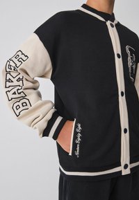 Chaqueta estilo varsity en negro y crema con botones a presión, puños acanalados y bordado en el pecho que dice "Baker". Tejido texturizado con mangas de contraste.