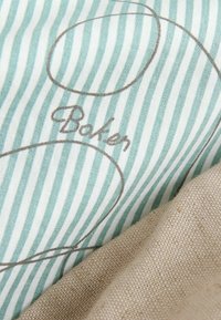 Tissu rayé avec des lignes bleu clair et blanches, comportant un texte cursif marron « Baker » et des contours ovales, partiellement recouvert d’un tissu beige texturé.