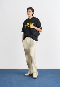 Melns pārmērīgs T-krekls ar dzelteno "Reebok" logo, bēšas sporta bikses ar tumšākiem traipiem un baltas kedas ar brūniem zolēm. Mazs melns somiņš.