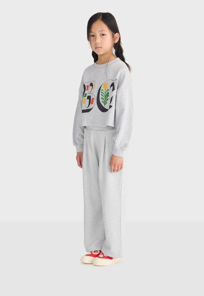 Jeune fille debout, portant un sweat-shirt gris clair avec des lettres florales "BG", un pantalon large assorti et des chaussures rouges à semelles blanches.