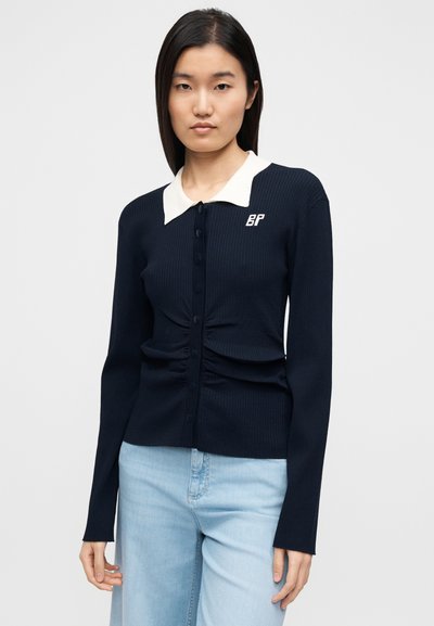 Μπλε ναυτικό ριμπ ζακέτα με λευκό γιακά, κουμπιά μπροστά και λεπτομέρειες σούρας στη μέση. Διαθέτει κεντημένο λογότυπο στο στήθος.