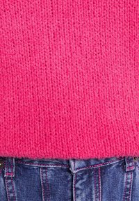 Helles, pinkes Strickpullover mit einem strukturierten Muster, kombiniert mit blauen Jeans, die an der Taille kontrastierende rosa Nähte aufweisen.