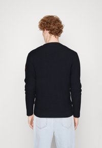 Granatowy sweter dzianinowy z prążkowaną fakturą, okrągłym dekoltem i długimi rękawami. Posiada logo na lewej stronie ramienia. Noszony z jasnoniebieskimi dżinsami.