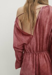 Robe en lin rouge profond à manches longues, avec un dos ouvert et une taille froncée, présentant une coupe décontractée et de fines bretelles.