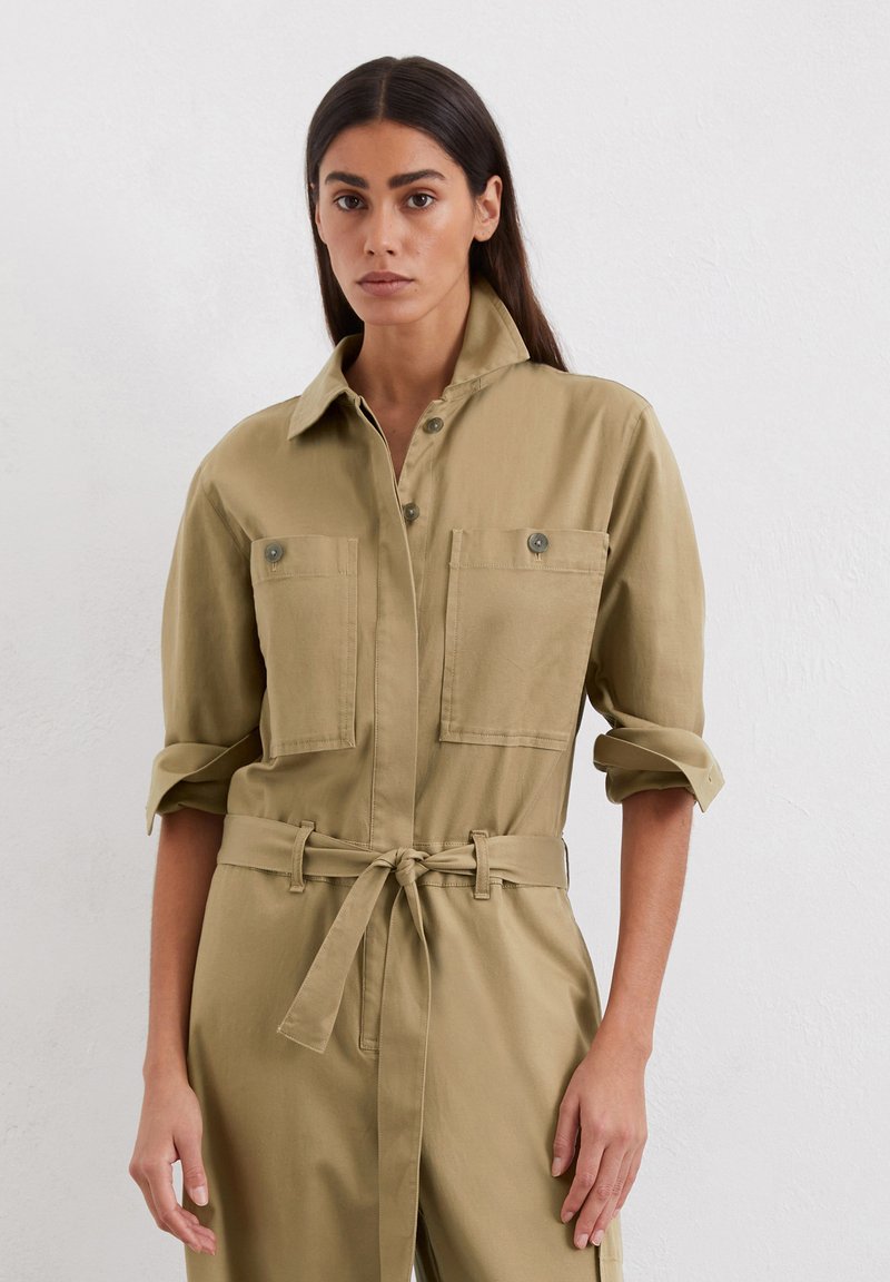 Hellbrauner Utility-Overall mit zwei Brusttaschen, Knopfverschluss, hochgekrempelten Ärmeln und einem gebundenen Taillengürtel. Glatte Stoffstruktur.