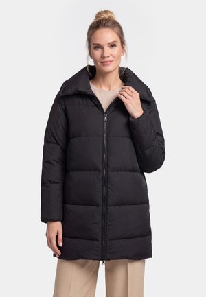 Frau mit blondem Haar im Dutt, trägt eine schwarze gesteppte Daunenjacke und beige Hose, steht vor einem einfarbigen hellen Hintergrund.