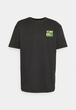 Homme portant un t-shirt graphique noir, un pantalon cargo foncé, des baskets blanches et des chaînes argentées sur un fond blanc uni.