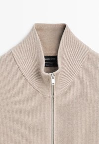 Beżowy bawełniany sweter z prążkowanym wzorem, zapinany na suwak, z wysokim kołnierzem, ozdobiony srebrnym zamkiem błyskawicznym i miękką, fakturowaną powierzchnią.