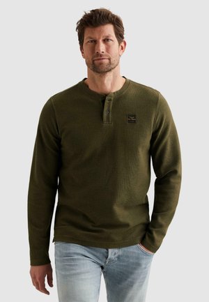 Man draagt een olijfgroen lang mouw henley-shirt met knoopsluiting en lichtblauwe spijkerbroek, staand met één hand in zijn zak.