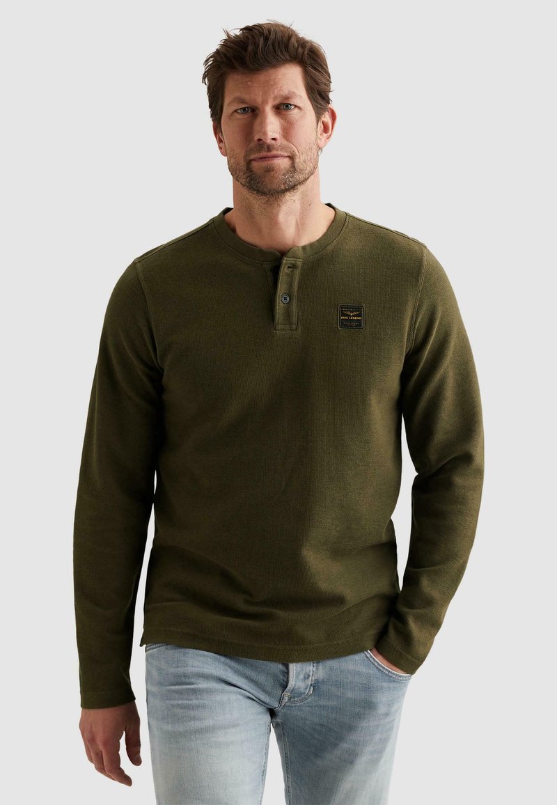 Homme portant une chemise henley à manches longues vert olive avec patte de boutonnage et un jean bleu clair, debout avec une main dans la poche.