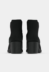 Par de botas negras por el tobillo con tacones gruesos, vistas desde atrás sobre un fondo blanco.