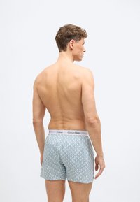 Άνδρας με κοντά, σγουρά μαλλιά, γυμνή πλάτη που γυρίζει με την πλάτη, φοράει ανοιχτό μπλε boxer Calvin Klein με σχέδια, πάνω σε απλό φωτεινό φόντο.