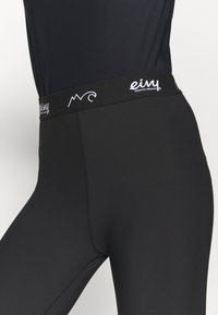 Leggings neri con un design aderente, dotati di una cintura larga nera contrassegnata da un marchio bianco, texture liscia e cuciture piatte.