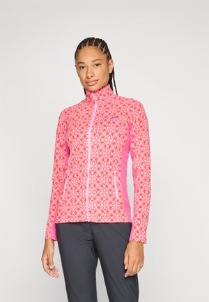 Donna che indossa una giacca rosa con motivo e zip, con pannelli laterali, in piedi davanti a uno sfondo chiaro e uniforme.