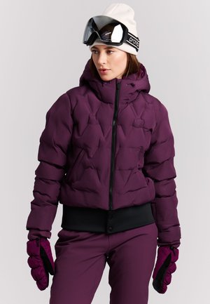 Vrouw draagt een paarse gewatteerde winterjas, bijpassende handschoenen en broek, een witte muts en een zwarte skibril op haar hoofd.