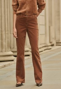 Pantalons en velours côtelé marron évasés avec une fermeture à bouton, dotés de poches latérales et d'une texture lisse, associés à un pull en laine marron doux.