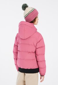 Giacca invernale rosa con cappuccio, leggera, design trapuntato e orlo a costine neri. Abbinata a un berretto lavorato a maglia a righe beige e rosa.