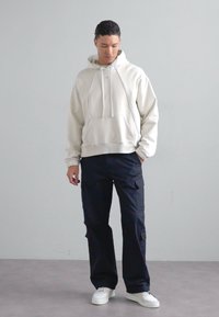 Hanorac bej deschis cu buzunar tip cangur, purtat împreună cu pantaloni cargo navy închis, dotati cu buzunare laterale, complectați cu adidași albi.