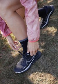 Zwarte Gore-Tex trailschoenen hebben een textuur bovenkant met veters, een contrasterende witte streep en een robuuste zool, geplaatst op gras met een hand die ze aanpast.