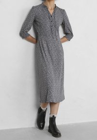 Lång marinblå klänning med vitt blommönster, figursydd midja, v-ringning och trekvartslånga ärmar, som bärs med svarta ankelskor.