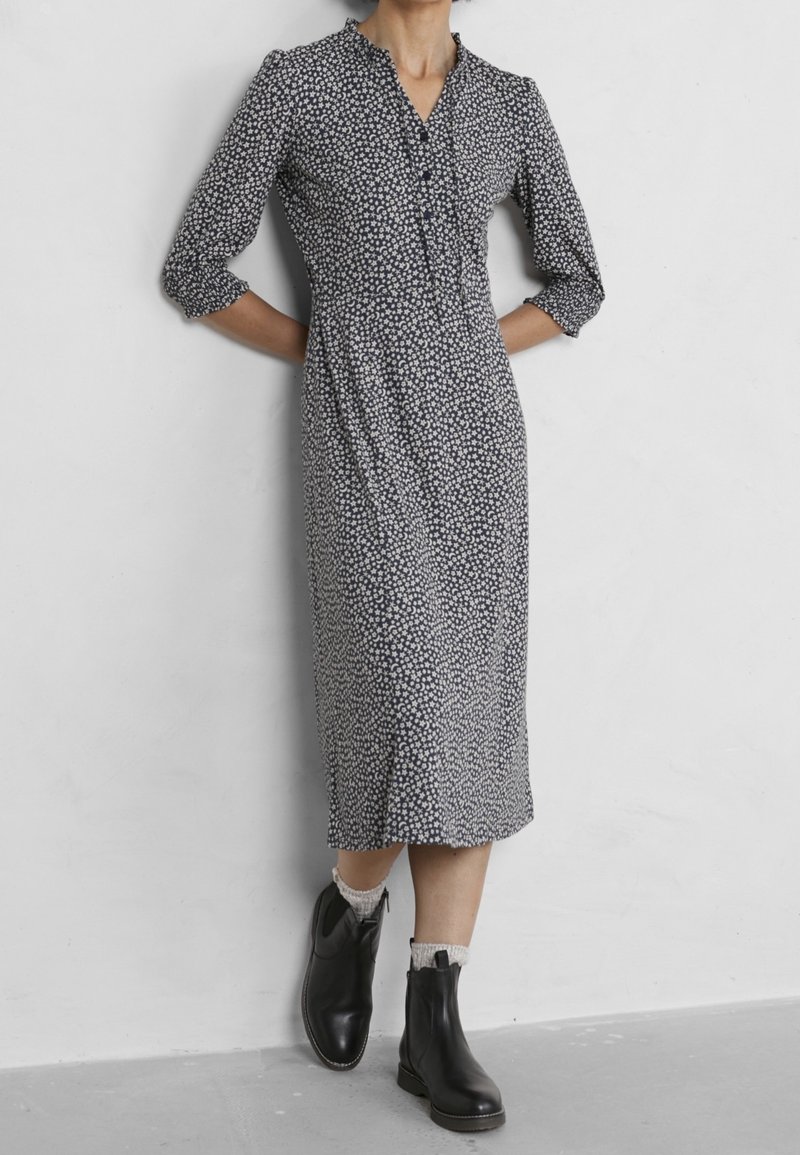 Lång marinblå klänning med vitt blommönster, figursydd midja, v-ringning och trekvartslånga ärmar, som bärs med svarta ankelskor.