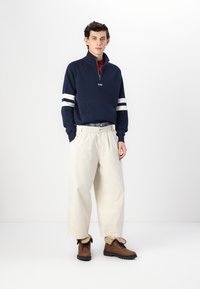 Marineblå halv-zip genser med hvite striper, lysebeige løstsittende bukser og brune snørestøvler. Materialet ser mykt og solid ut.