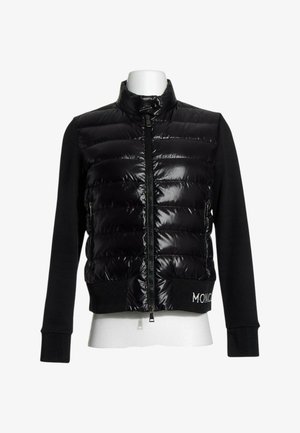 Chaqueta acolchada negra con cremallera frontal, mangas largas de tela y puños de canalé, expuesta en un maniquí blanco sin cabeza.