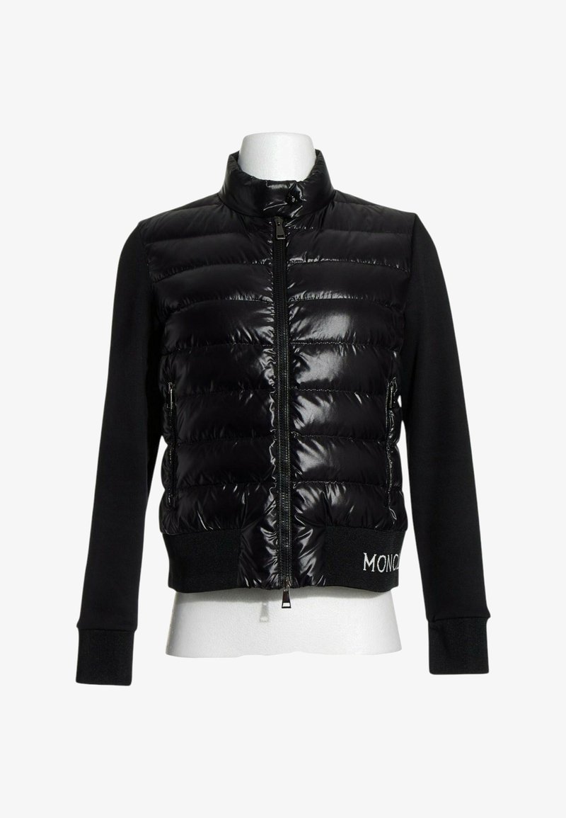 Veste matelassée noire avec fermeture éclair à l'avant, manches longues en tissu et poignets côtelés, présentée sur un mannequin blanc sans tête.