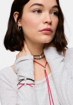 Joven mujer con pelo corto y castaño que lleva una camiseta gris claro y collares y pulseras de cuerda rojos y negros con pequeños cierres plateados.