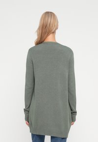 Cardigan deschis, cu mânecă lungă, din tricot verde. Are o croială lejeră, ba chiar și un tiv și manșete ribbate, cu un aspect neted.