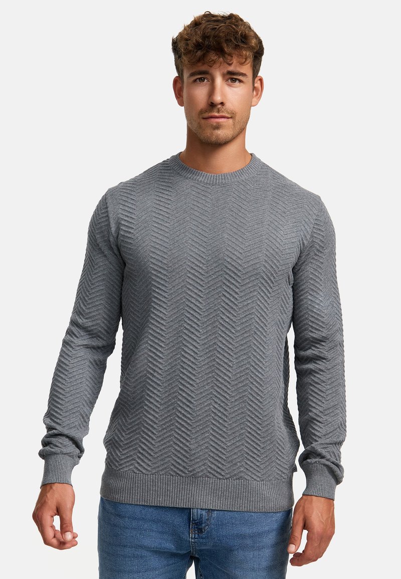 Pulover gri tricotat cu un model texturat de herringbone, decolteu rotund și manșete și tiv cu dungi. Combinat cu blugi albaștri.