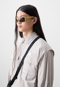 Sunglasses with brown lenses and light wood-patterned frames featuring a green logo accent, worn with a light gray button-up shirt and strap.