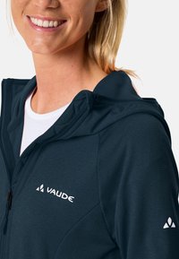 Felpa Vaude di colore blu scuro con zip frontale, tessuto leggero e logo sul petto e sulla manica. Presenta un cappuccio e materiale testurizzato.
