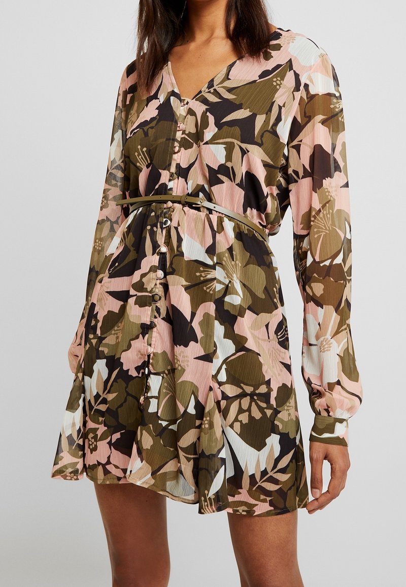 Femme portant une robe boutonnée à manches longues avec un motif floral et une fine ceinture à la taille, sur un fond neutre.