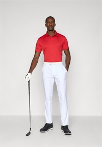 Camisa polo vermelha de manga curta, com detalhes texturizados; combinada com calças brancas ajustadas e sapatos de golfe pretos. Segurando um taco de golfe.