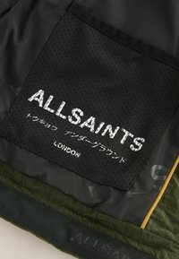 Juodas audinio pleistras su baltu tekstu "ALLSAINTS" ir "LONDON", su tekstūruotu fonu ir žaliu audiniu apačioje.