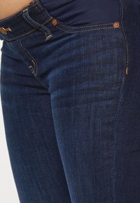 Jeans de denim azul escuro com um design ajustado. Apresenta costuras visíveis, acessórios metálicos e uma superfície suavemente texturizada.