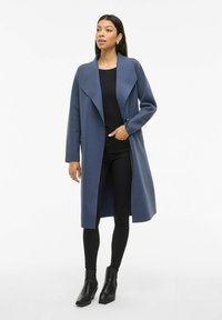 Lungo cappotto blu con ampio colletto, realizzato in tessuto liscio, dotato di tasche laterali, indossato sopra una maglia nera e pantaloni, abbinato a stivaletti neri.