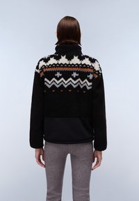 Persona con capelli castani lisci indossa una giacca in pile nera con motivi geometrici bianchi, arancioni e grigi, abbinata a pantaloni grigi, di spalle.