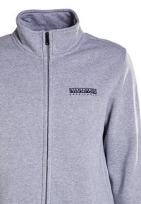 Giacca zip-up grigia chiara realizzata in tessuto morbido, con colletto alto e un logo ricamato nero sul lato sinistro del petto.