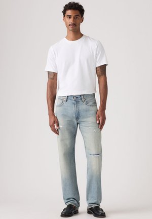 Homme aux cheveux courts bouclés et tatouages, portant un t-shirt blanc uni, un jean bleu clair déchiré et des mocassins noirs, debout devant un fond neutre.