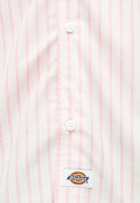 Λευκό ύφασμα με κατακόρυφες ροζ ρίγες, δύο λευκά κουμπιά στο κέντρο και ένα ορθογώνιο έμβλημα με το λογότυπο Dickies κοντά στο κάτω μέρος.