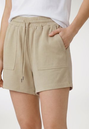 Beżowe casualowe szorty z elastycznym paskiem, sznurkiem i dużymi przednimi naszywanymi kieszeniami, noszone z włożoną białą bluzką.