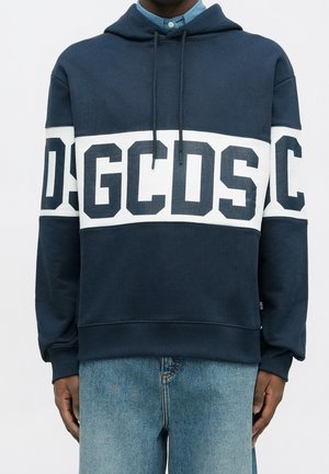 Personne portant un sweat à capuche bleu marine avec une bande horizontale blanche et de grandes lettres sombres GCDS sur la poitrine et les manches, associé à un jean bleu clair.