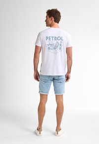 Bílý tričko s modrou grafikou "PETROL" a designem motocyklu na zádech, spárované s světle modrými zkrácenými džínovými šortkami a neutrálními teniskami.
