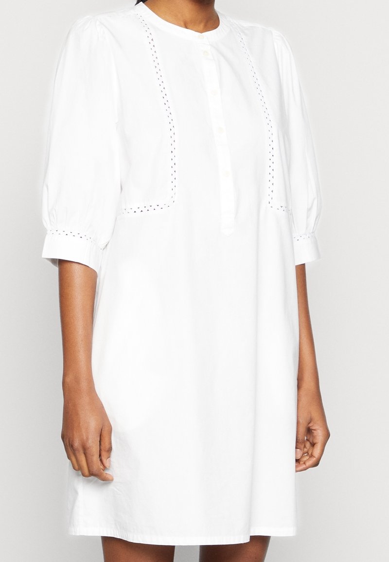 Femme portant une robe blanche mi-longue avec des manches ballon trois-quarts et des détails subtils en dentelle ajourée sur le devant et les poignets.