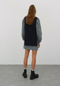 Marineblau gestrickte Weste über einem karierten Hemdkleid, schwarze klobige Stiefel und graue Sockenakzente, von hinten gegen einen schlichten Hintergrund betrachtet.